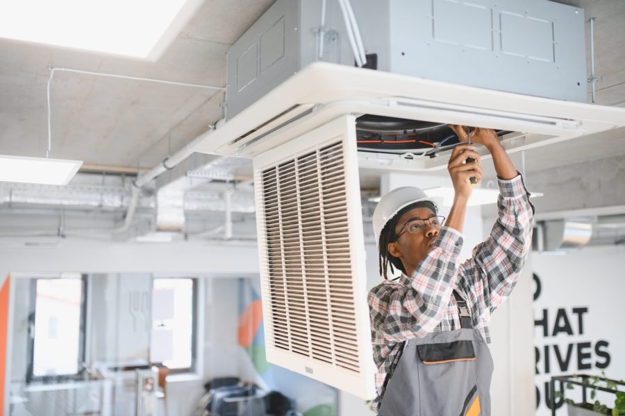 Technician in a hard hat and uniform working on an air conditioning unit in a modern office, ensuring proper ventilation and cooling