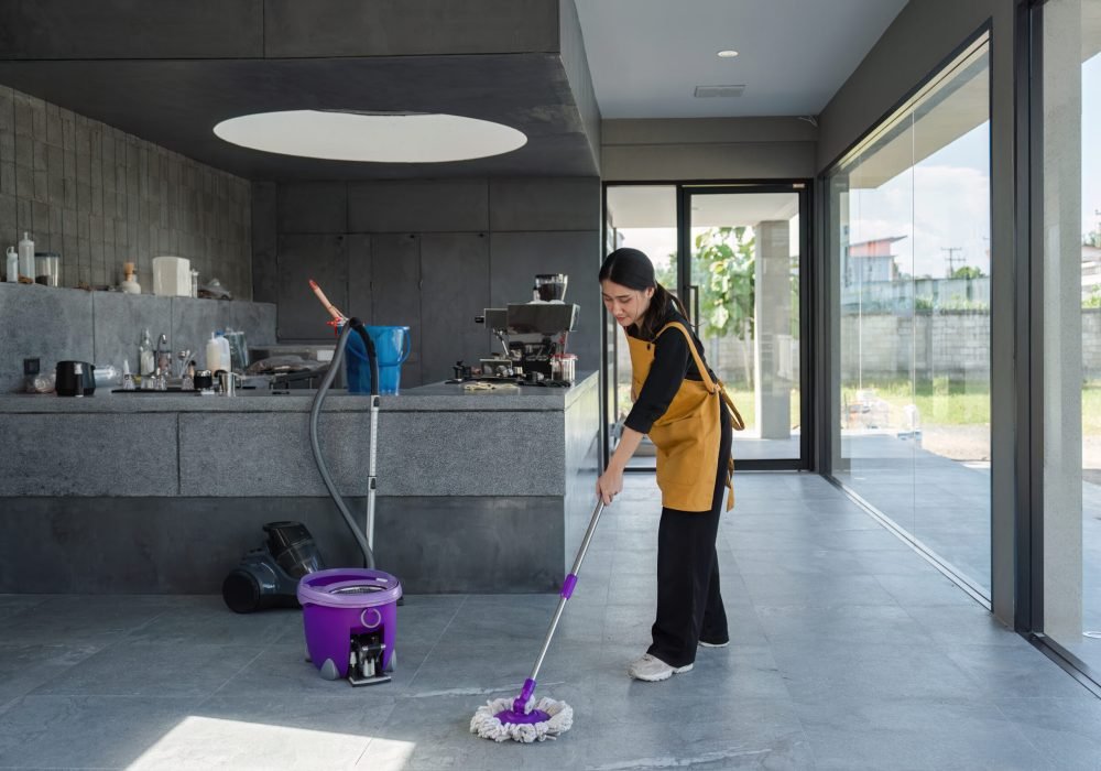 A female employee in a modern coffee shop cleans the floor with a mop and bucket, ensuring a tidy and welcoming environment.